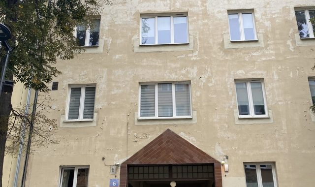 Elewacja budynku z przejściem. Budynek o jasnej, kremowej elewacji z widocznymi śladami zużycia. Na parterze centralnie umieszczone przejście bramowe z brązową podmurówką i daszkiem w kształcie trójkąta. Nad przejściem i po bokach rzędy prostokątnych okien. Po lewej stronie widoczne drzewa, po prawej stojaki na rowery.