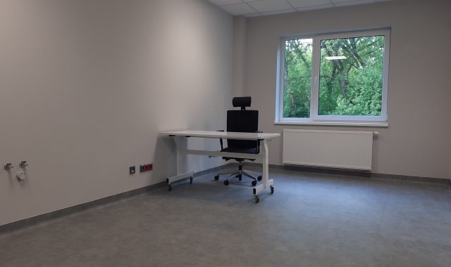 Po lewej stronie na dłuższej ścianie są widoczne nad podłogą oddalone od siebie dwa punkty z gniazdkami. Po lewej stronie na dłuższej ścianie widoczne jest biurko i krzesło