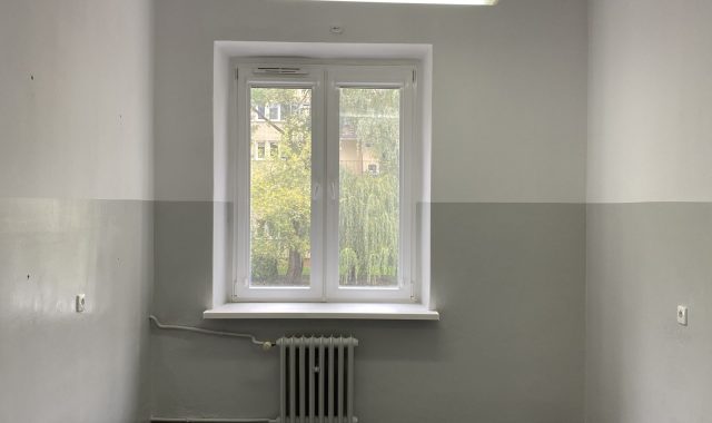 Wnętrze pustego pokoju z oknem. Prostokątne pomieszczenie z białymi ścianami i ciemnoszarą wykładziną na podłodze. Na krótszej ścianie znajduje się okno z białą ramą, pod nim grzejnik żeliwny. Na suficie podłużna lampa świetlówkowa. Pomieszczenie jest puste, bez mebli.