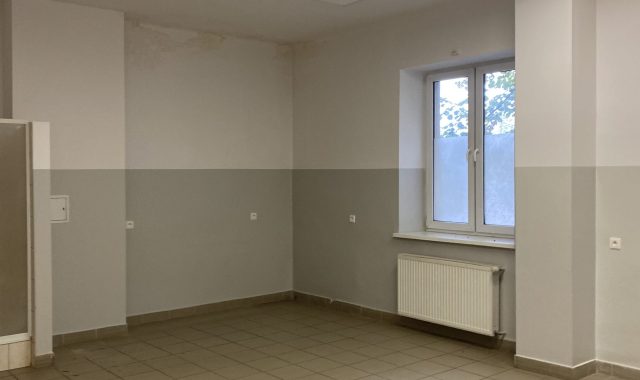 Pomieszczenie z płytkami na podłodze. Prostokątne pomieszczenie z jasnoszarymi ścianami i beżową podłogą z dużych płytek. Na krótszej ścianie okno z białą ramą, pod nim grzejnik panelowy. Na suficie kilka prostokątnych lamp LED.