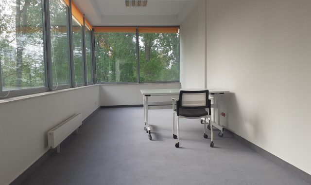 Po lewej stronie pod oknami jest grzejnik. Pośrodku pokoju przy prawej długiej ścianie widoczne są dwa biurka i krzesło na kółkach.