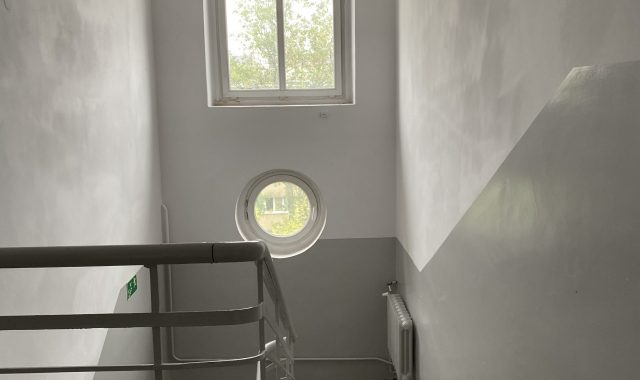 Klatka schodowa z oknem okrągłym. Wnętrze klatki schodowej z betonowymi schodami i białą metalową balustradą. Na ścianie okrągłe okno oraz prostokątne okno powyżej. Ściany i sufit pomalowane na biało i jasnoszaro, podłoga jasnoszara.