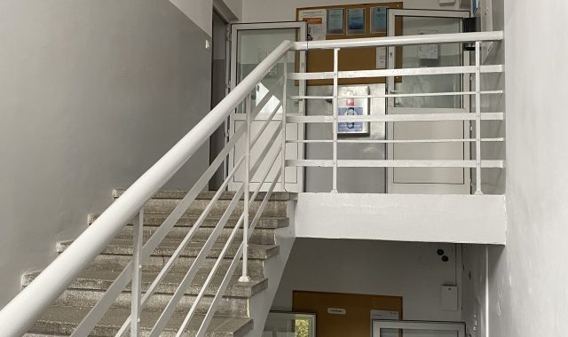 Ostatnie piętro z klatką schodową i tablicami. Widok z półpiętra na klatkę schodową z białą metalową balustradą. W tle ściana z tablicami informacyjnymi i gablotami. Podłoga jasnoszara, ściany białe, sufit z prostokątnymi lampami.