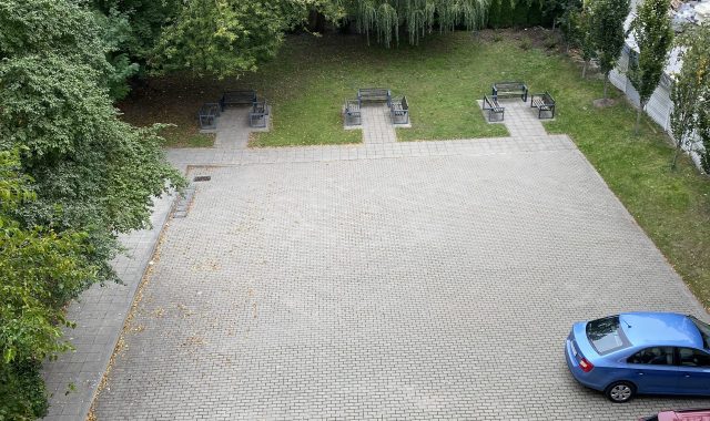 Parking z samochodami i zielenią. Utwardzony parking z kostki brukowej, na którym stoją dwa samochody: czerwony i niebieski. Po prawej stronie pas zieleni z drzewami i krzewami, na wprost ławki. W tle widoczne budynki mieszkalne.